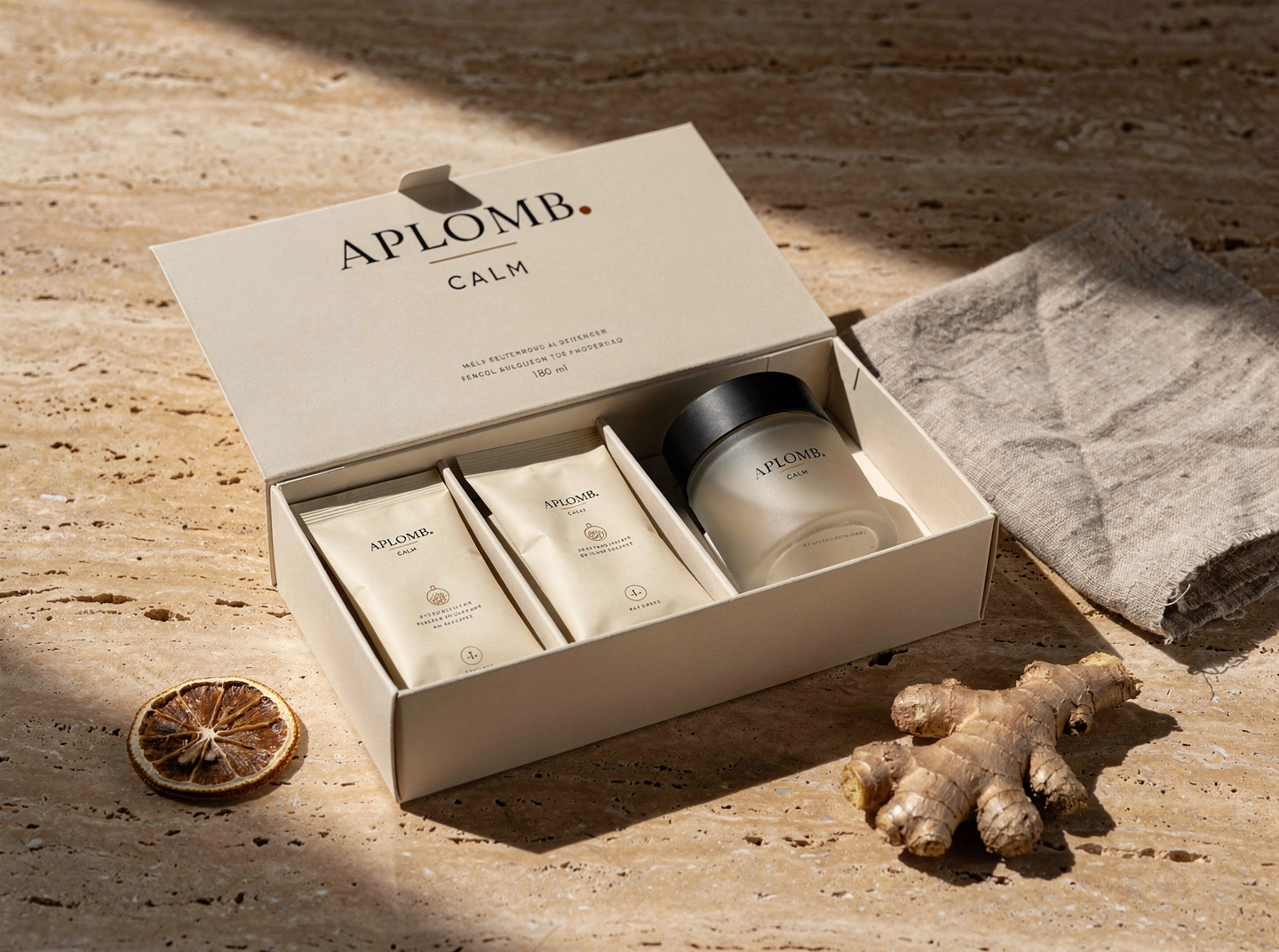 APLOMB. Calm — open cream paperboard kit-box reading 'APLOMB · CALM' containing herbal sachets, a small frosted-glass jar, and an instruction card; dried lemon and fresh ginger root beside it on travertine.