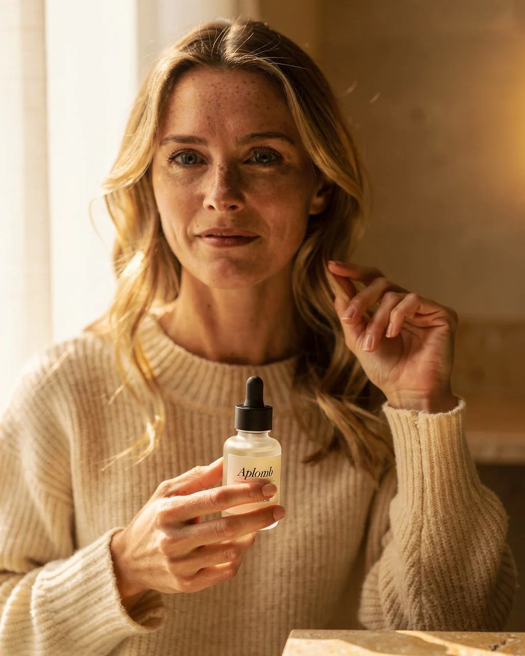 A woman in her thirties holding the APLOMB Facial Serum in warm window light.