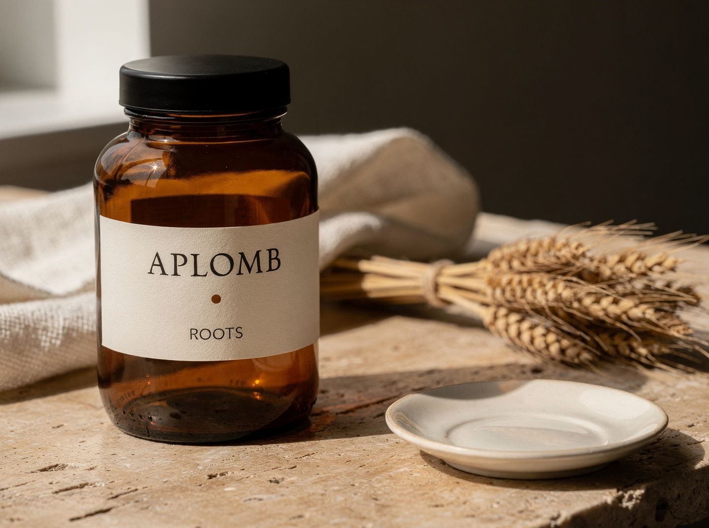 APLOMB. Roots — amber-glass apothecary jar with a cream paper label reading 'APLOMB · ROOTS' beside a small bundle of dried wheat stalks on travertine in warm window light.